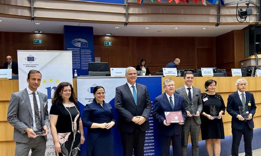 Ludzie stojący w rzędzie elegancko ubrani trzymający nagrody. W tle  sala Parlamentu Europejskiego